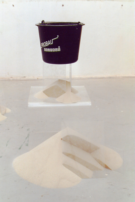 Skulptur aus Sand mit darinsteckendem Plexiglas, darüber ein Eimer
